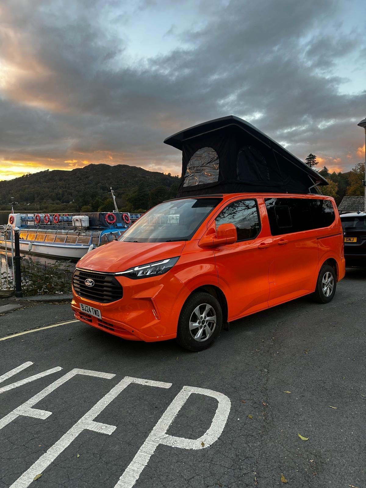 Orange JMC pop-top campervan with roof raised at a lakeside at sunset